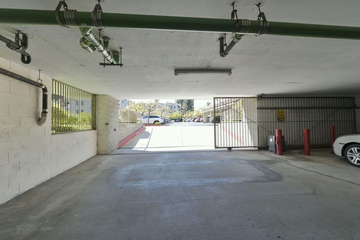 A view from inside an underground parking garage, showing an exit with a gated entrance. Green pipes run along the ceiling, and there are red poles marking the area. Outside, a paved area with parked cars and greenery can be seen in the distance. Natural light illuminates the scene.