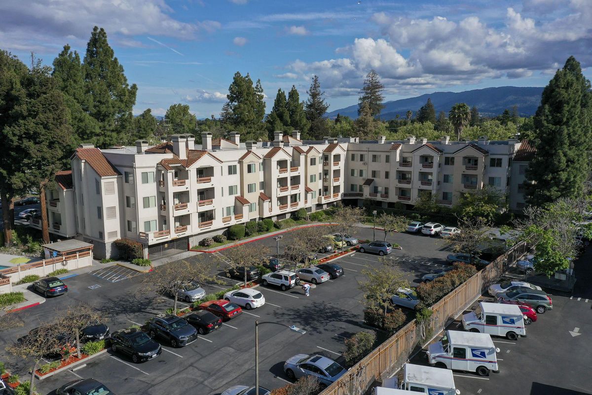 Aerial view of a multi-story residential building surrounded by trees and a parking lot filled with cars. The sky is partly cloudy, and mountains are visible in the background.