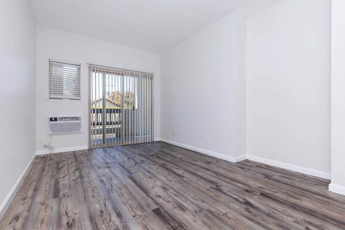 Empty room with light gray walls and wooden flooring. There are vertical blinds covering a large window that opens to a balcony. An air conditioning unit is mounted on the wall near the window. The space is bright and airy, with ample natural light coming in.