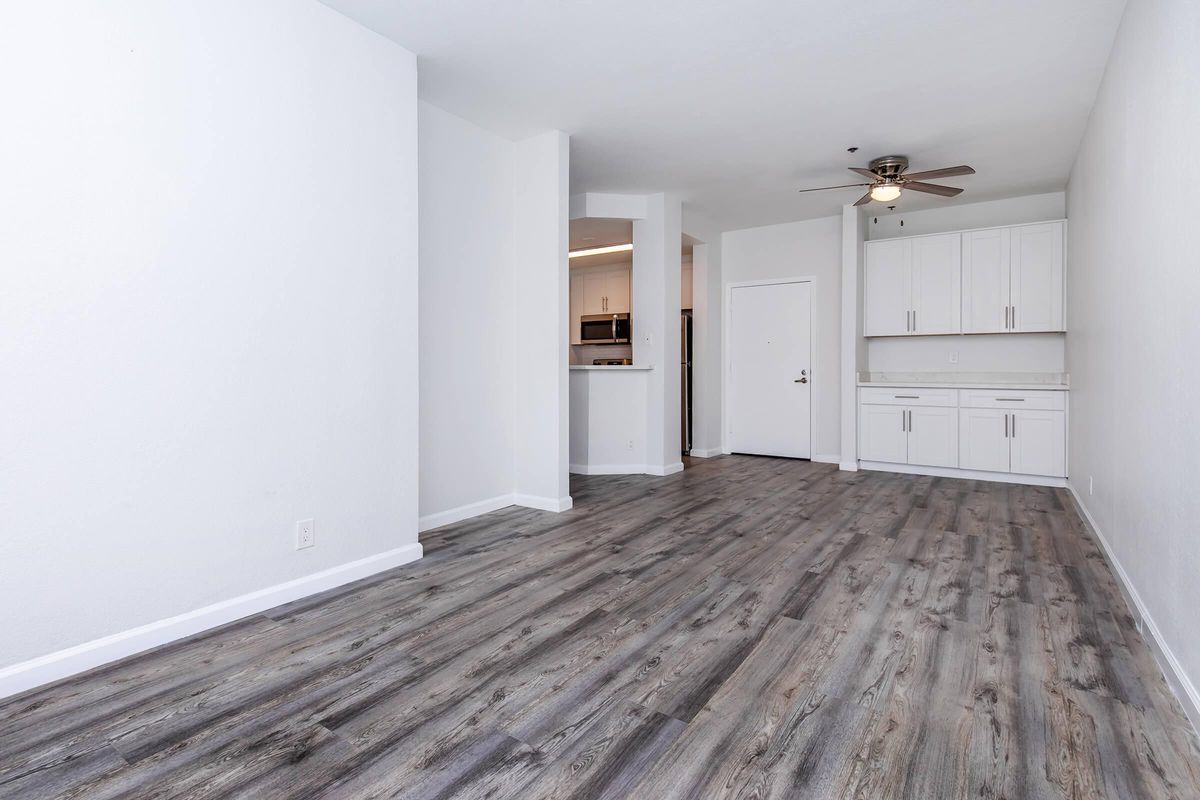 Spacious, modern open-concept living area featuring light-colored walls, laminate flooring, and a ceiling fan. The kitchen is partially visible with white cabinetry and stainless steel appliances. Natural light fills the room, creating a welcoming atmosphere.