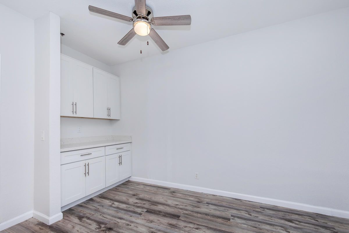 An empty room featuring light-colored walls, a ceiling fan with wooden blades, and a white cabinet with countertop. The floor is made of wood-like planks, creating a spacious and modern atmosphere. The room is well-lit, emphasizing its uncluttered design.
