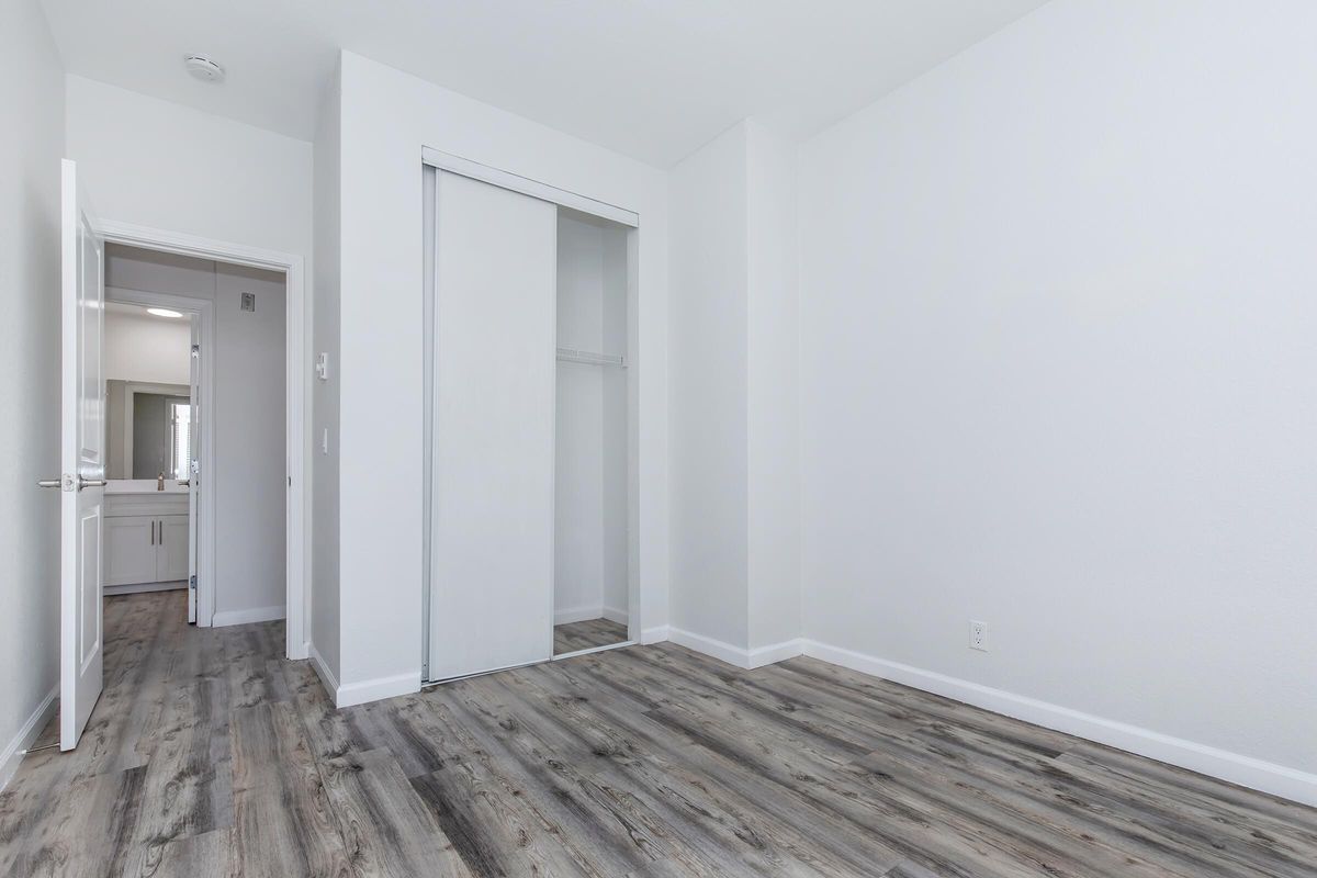 A bright, empty room with white walls and a light wood floor. There is a mirrored closet door on the left. A doorway leads to another room, partially visible, which appears to include a bathroom area. The overall atmosphere is clean and minimalistic.