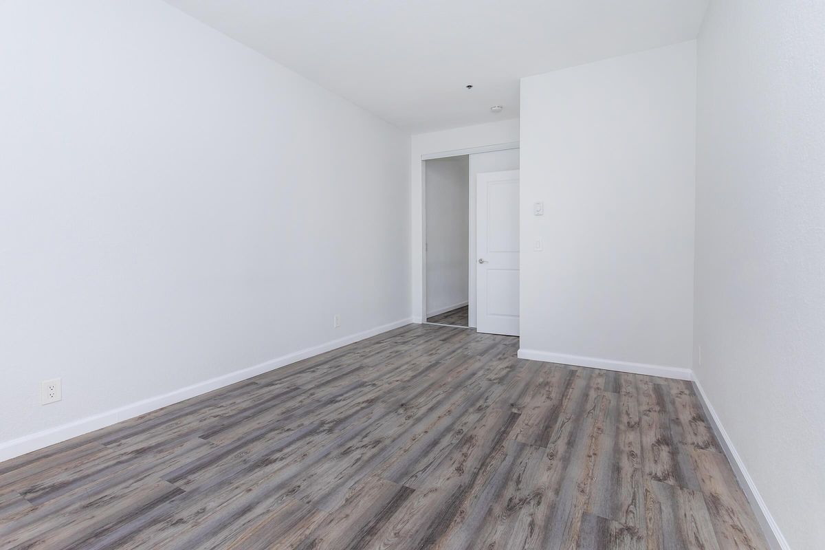 Empty room with light gray walls and wood-like flooring. A door is visible on the left side leading to another room. The space is well-lit, giving a clean and modern appearance, suitable for various uses such as a living area or office.