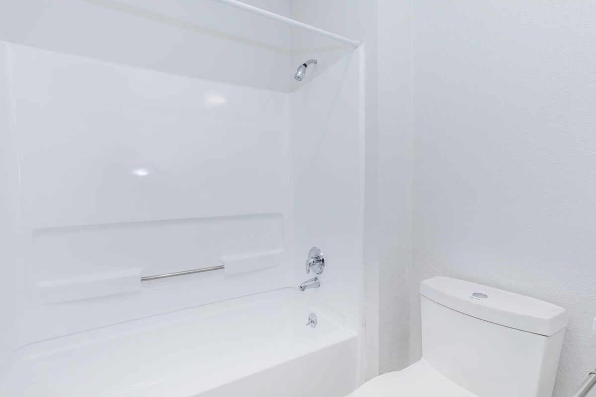 A clean, modern bathroom featuring a white bathtub with a showerhead, a silver grab bar, and a white toilet. The walls are painted a light color, creating a bright and spacious atmosphere.