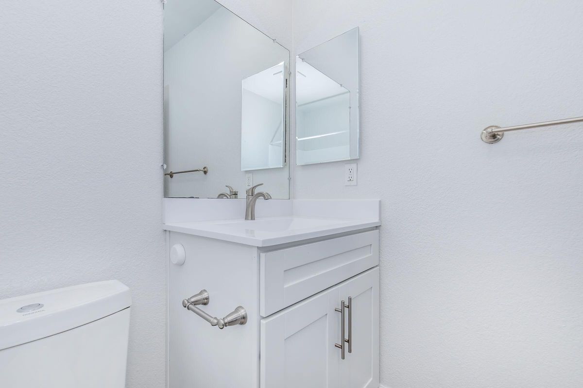 A clean, modern bathroom featuring a white vanity with a sink and a wide mirror above it. There is a toilet visible on the left side of the image. The walls are painted white, and the space appears bright and well-lit. A towel rack is mounted on the right wall.