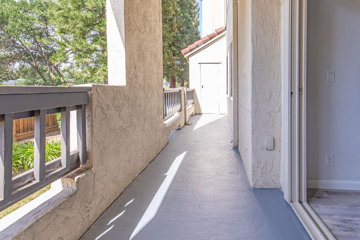 A bright, partially shaded corridor with a smooth concrete floor, flanked by textured walls. To one side, there is a railing overlooking greenery and light filtering through trees. The corridor leads to a white door at the far end, with natural light enhancing the space's openness.