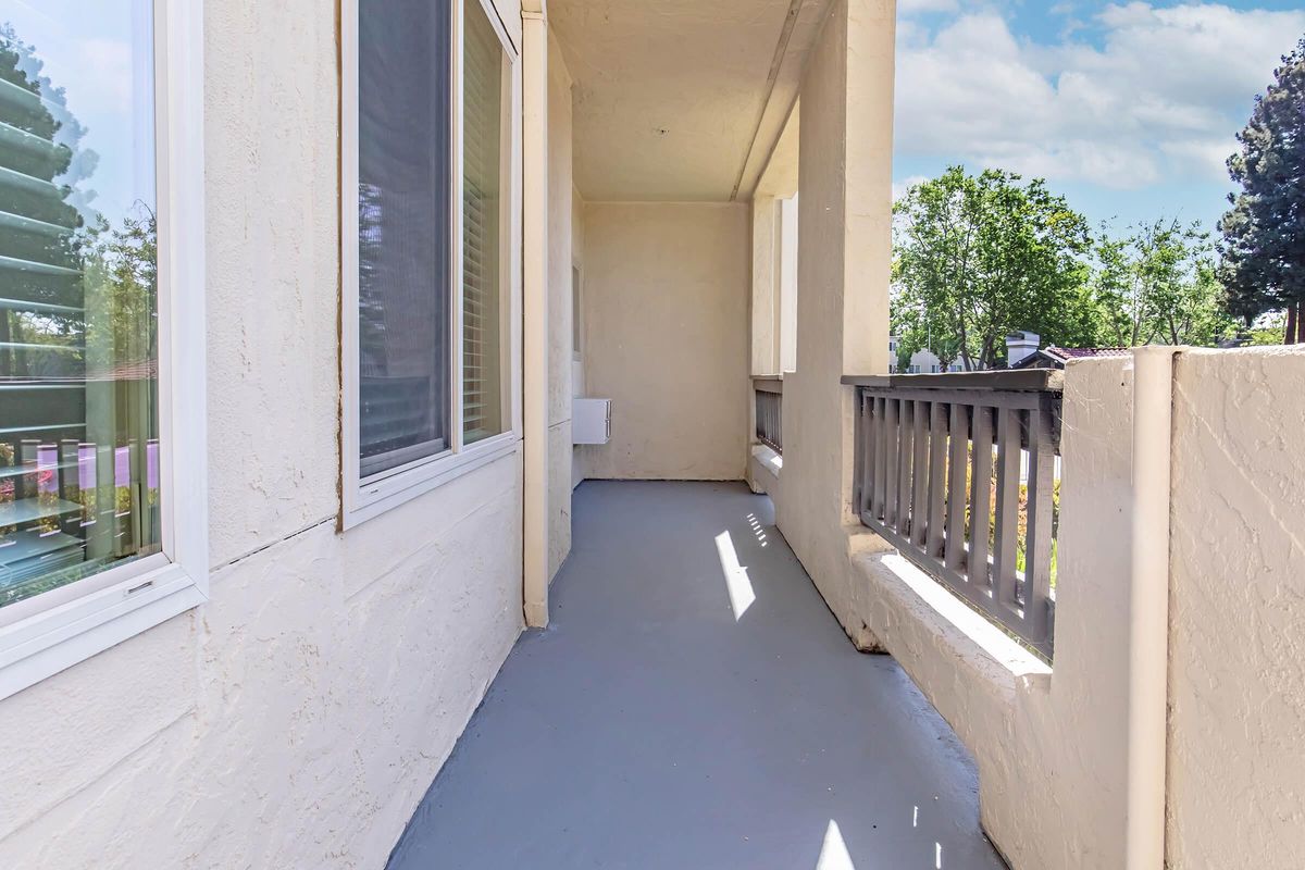 A clean, empty balcony with a light gray floor and white walls. Sunlight filters in through nearby windows, casting soft shadows. The railing is made of wood, and there are green trees in the background, contributing to a serene outdoor atmosphere.