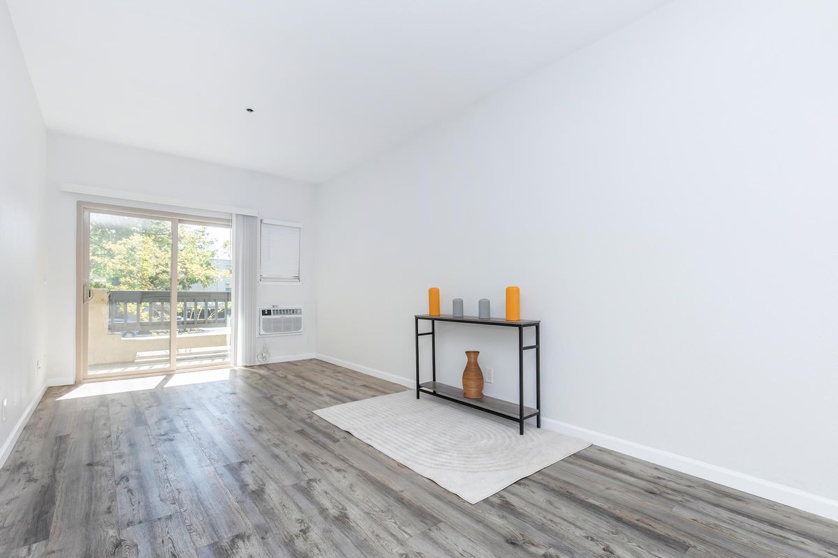A bright, empty living room featuring light wood flooring and white walls. A simple black console table holds three orange candles and a brown vase. In the background, there is a sliding glass door leading to a balcony with greenery visible outside. Natural light streams in, creating a spacious feel.