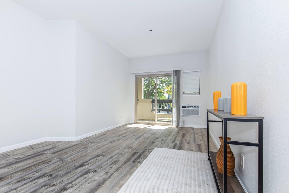 Bright, spacious interior of a living room featuring light wood-like flooring, white walls, and a sliding glass door leading to a balcony. A modern console table with orange decorative vases is positioned against the wall. Natural light fills the room, creating an inviting atmosphere.