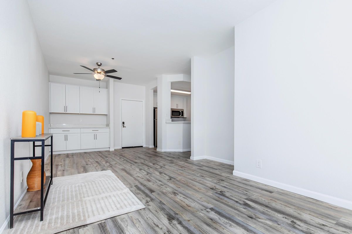 A bright and modern living space featuring light-colored walls and wood-like flooring. A small black console table with decorative items is to the left, and a kitchen area with white cabinets is visible in the background. A ceiling fan hangs above the room, creating a spacious and inviting atmosphere.