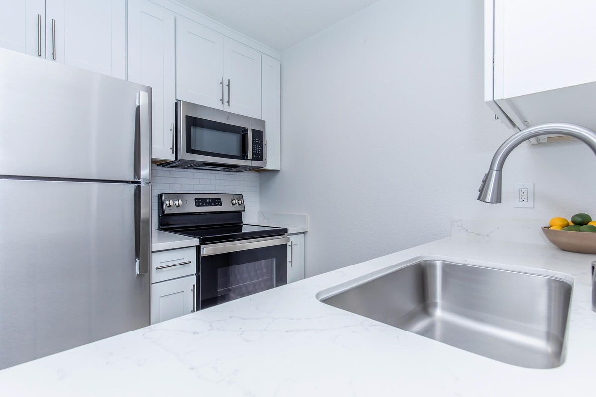 Modern kitchen featuring stainless steel appliances, including a refrigerator, microwave, and oven. The countertop is white with a marble-like finish, and a stainless steel sink is visible. Light-colored cabinets line the wall, contributing to a bright and clean aesthetic. A bowl of fruit is placed on the counter.