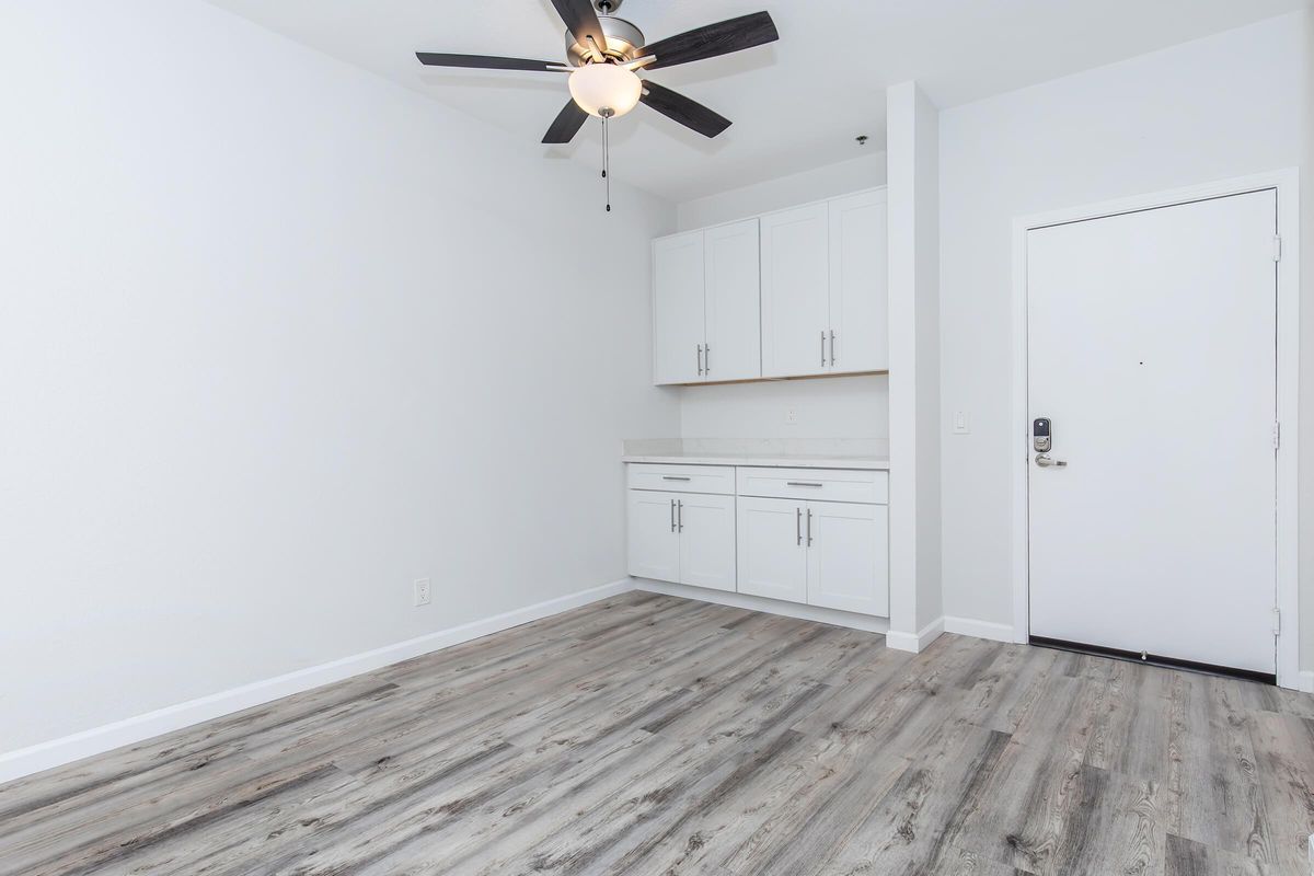 A spacious, empty room with light wood flooring, a ceiling fan, and white walls. It features white cabinetry along one wall and a door leading outside. Natural light enhances the airy feel of the space.