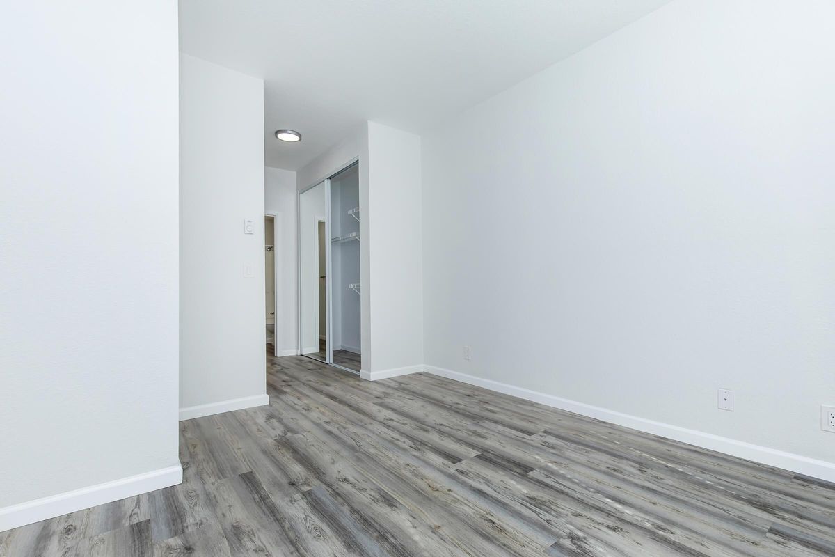 A spacious, empty room with light-colored walls and wooden flooring. The room features a closet area and an adjacent doorway leading to another room. Natural light illuminates the space, creating a bright and airy atmosphere.