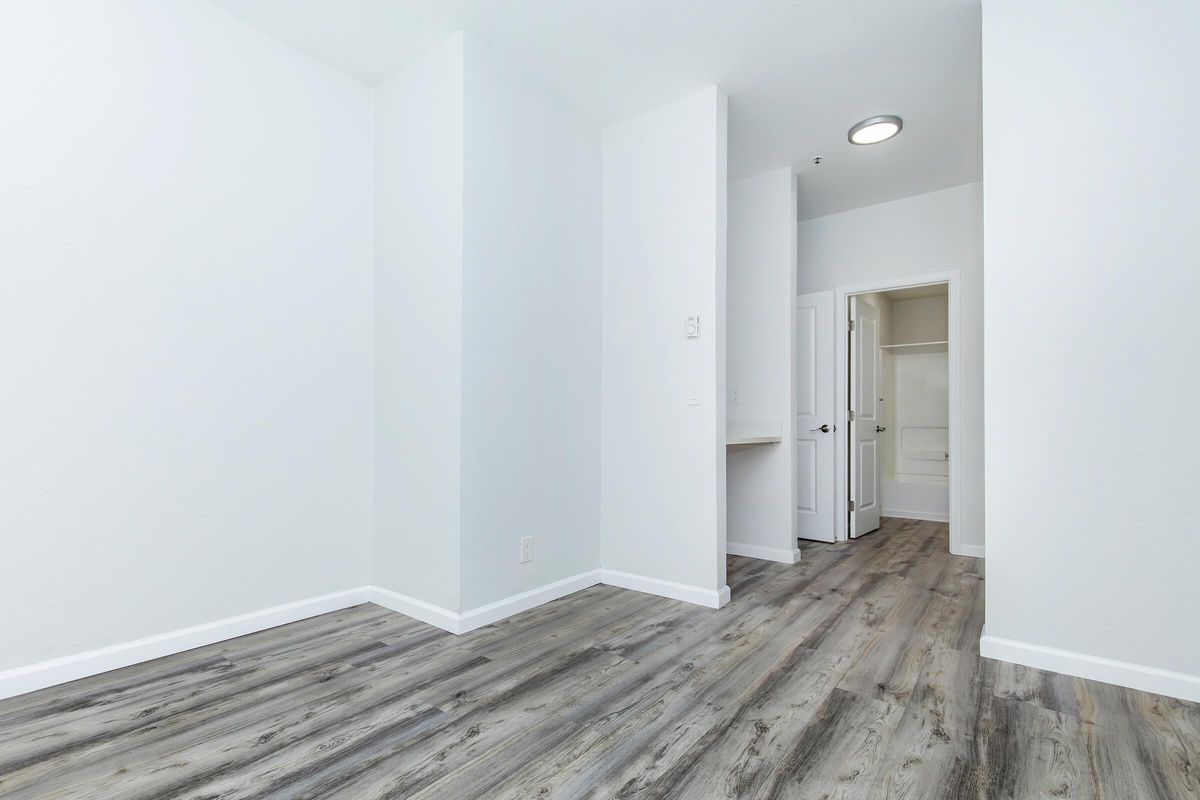 A bright, empty room with light-colored walls and a wood-like floor. There is a doorway on the right leading to a bathroom, and a small closet space visible. The ceiling has a round light fixture, and the overall atmosphere is clean and modern, suitable for various uses or furnishings.