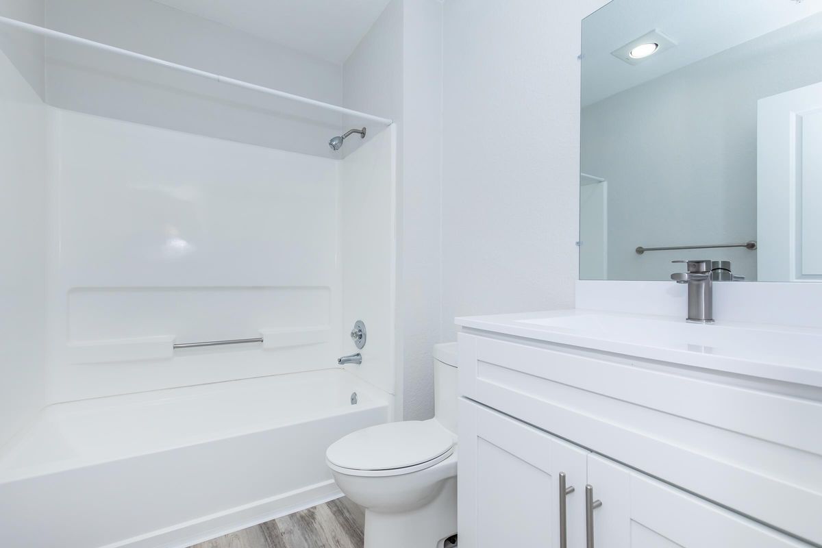 A clean, modern bathroom featuring a white bathtub with a shower, a wall-mounted mirror above a sleek white vanity with a sink, and neutral-colored walls. The flooring is light wood, enhancing the bright and fresh atmosphere of the space.