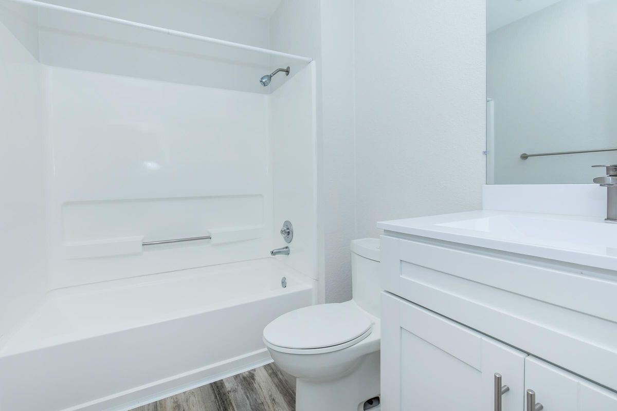 A clean, modern bathroom featuring a white bathtub with a showerhead, a white toilet, and a sleek vanity with a sink. The walls are painted light, and the flooring is a wood-like material, creating a bright and airy atmosphere.