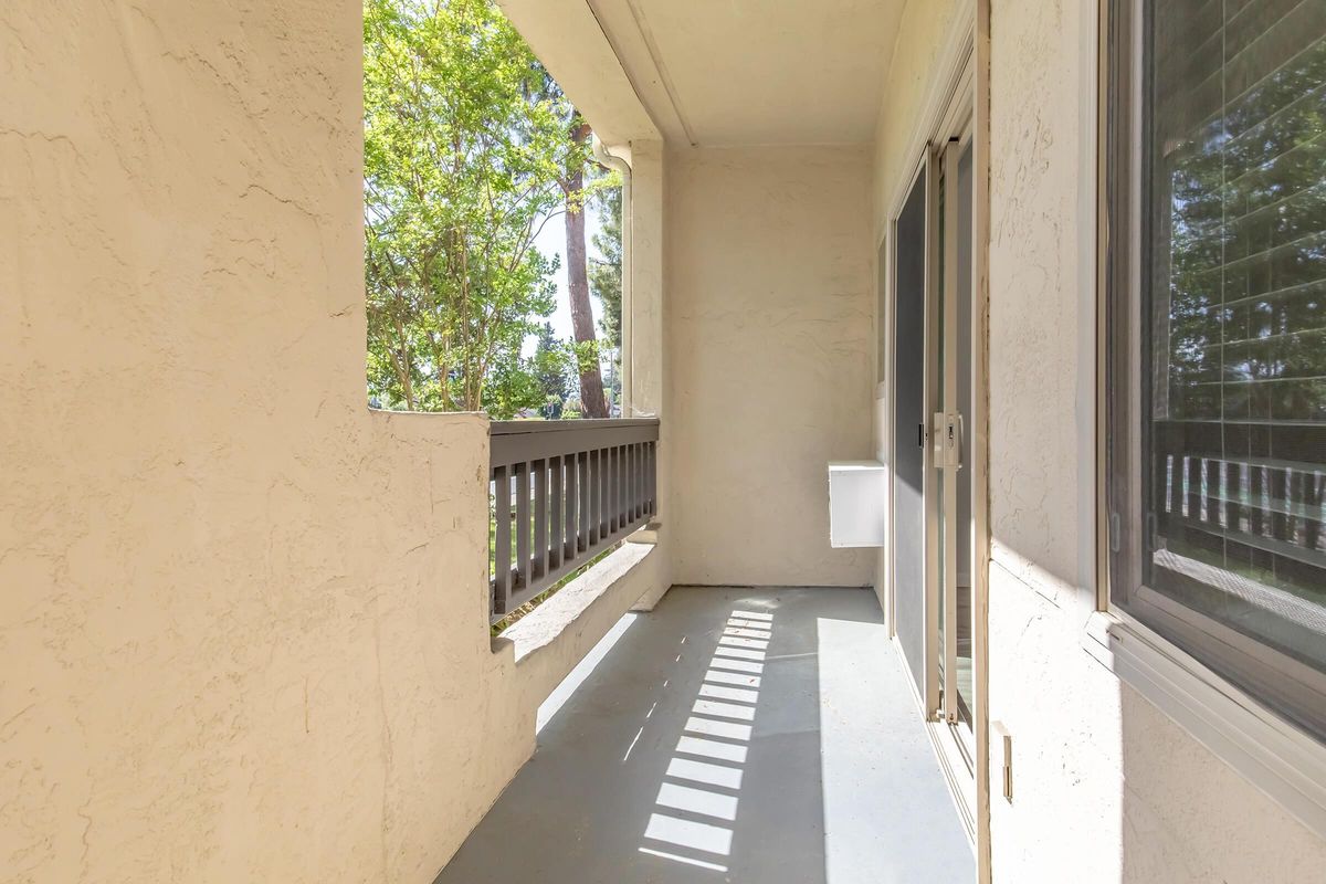 A small balcony with a light-colored surface, bordered by a low railing. Sunlight casts shadows from the railing onto the floor, and there are trees visible in the background, creating a serene outdoor space. The balcony is empty and offers a view of the surrounding greenery.