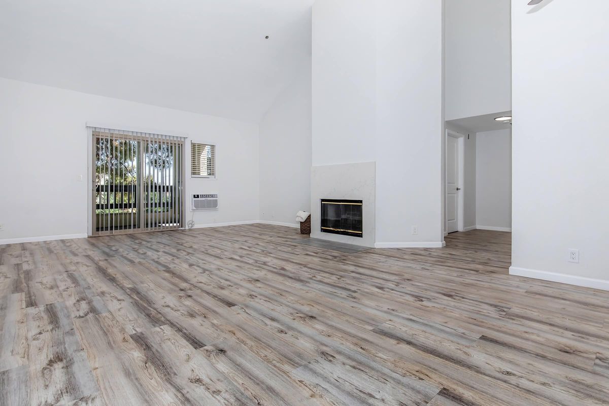 A spacious living room with light-colored walls and wood-style flooring. It features a white fireplace on one side, tall ceilings, and a large window letting in natural light. Vertical blinds cover the window, and there's an air conditioning unit mounted on the wall. The room is empty, creating an open and inviting atmosphere.