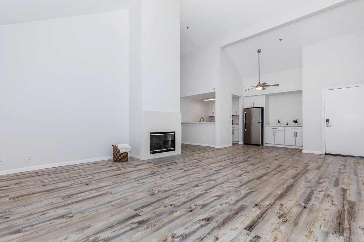 A modern, open-concept living space with light wood flooring, a white fireplace, and a ceiling fan. The kitchen area features stainless steel appliances and white cabinetry. The high ceiling and large windows create a bright atmosphere. A door leads to the entrance, offering a view of the living room.