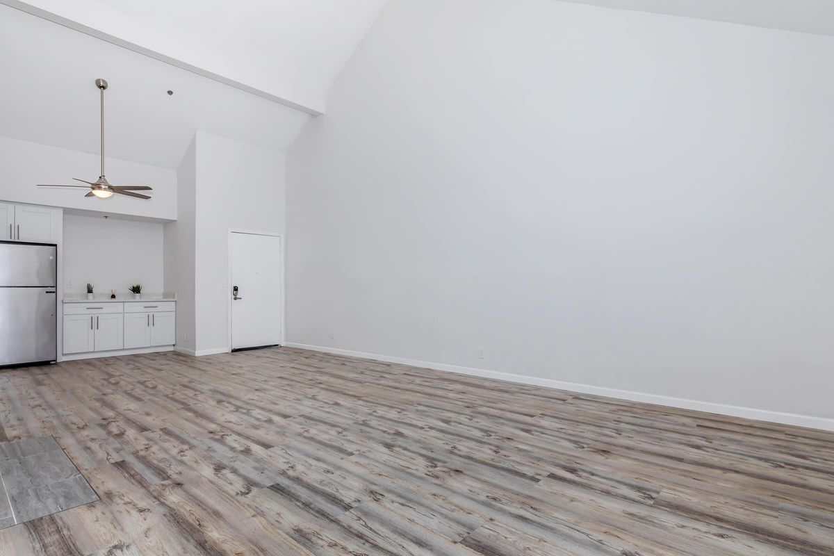 Spacious, minimalist living area with light-colored wooden flooring and high ceilings. A kitchen area with a stainless steel refrigerator is visible in the background. The walls are painted white, creating a bright, airy feel. A single door leads outside, enhancing the open layout.