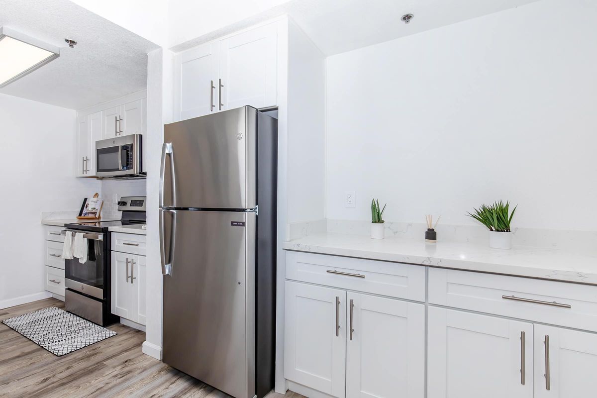 Modern kitchen featuring stainless steel appliances, including a refrigerator and microwave. White cabinets and light countertops create a bright atmosphere, complemented by a small decorative plant on the countertop. A patterned mat is visible on the floor, adding a touch of comfort to the space.