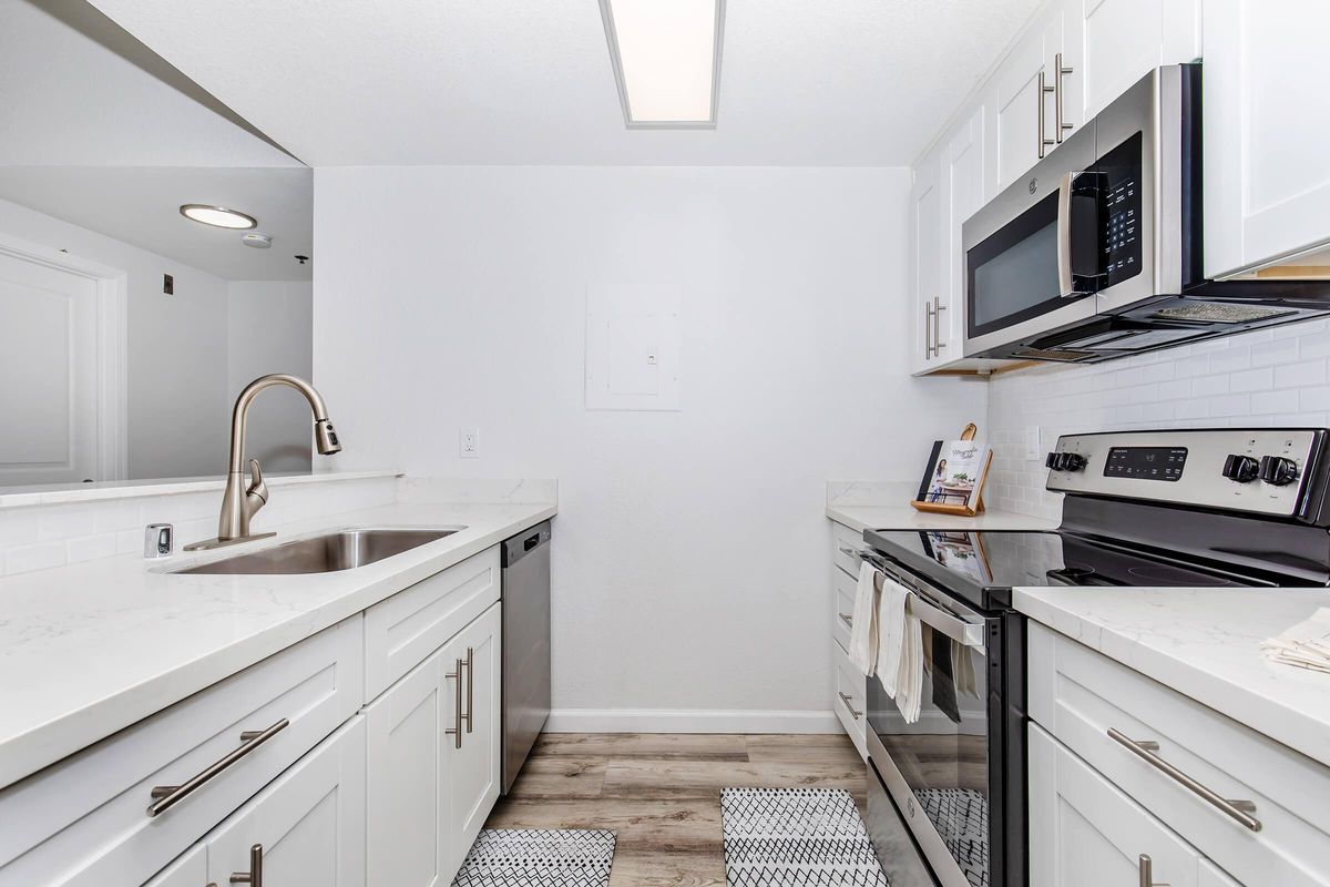 Modern kitchen featuring white cabinetry, stainless steel appliances including an electric stove and microwave, a stainless steel sink, and light-colored countertops. The floor has a wood-like finish, and there are decorative mats in front of the sink and stove. The space is bright and airy with neutral tones.