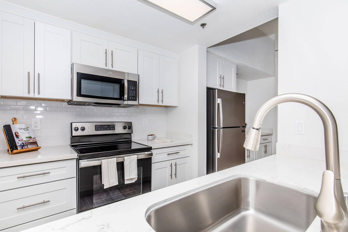 A modern kitchen featuring white cabinetry, a stainless steel microwave, and an oven. There is a double-basin sink with a stylish faucet in the foreground, and a refrigerator in the background. The countertops are made of a light-colored stone, and there's a small decorative item on the countertop.
