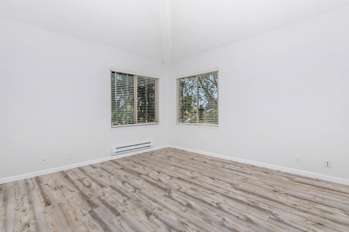 A bright, empty room with white walls and two large windows featuring blinds. The flooring is light wood, and the ceiling has a vaulted design. Natural light floods the space, casting soft shadows on the floor.