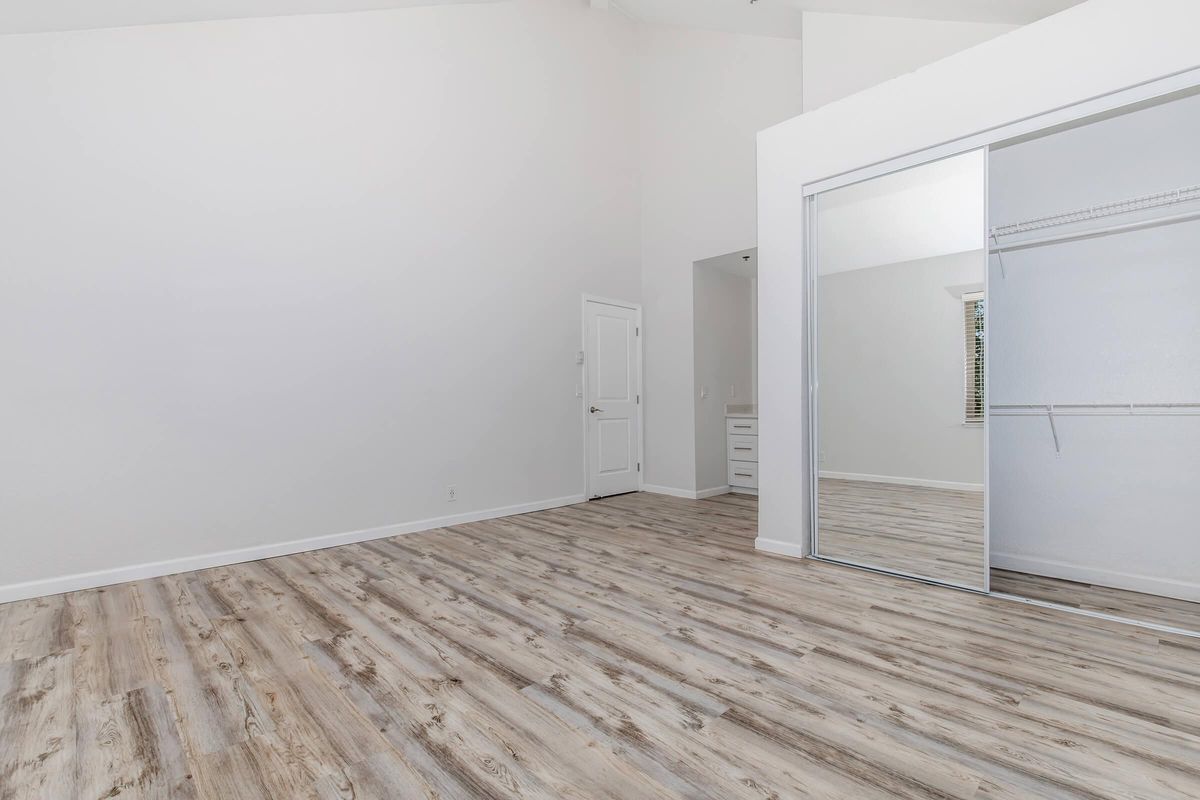 A spacious, empty room with high ceilings and a light-colored wall. Features include a wooden floor, a large mirrored sliding closet door, and a doorway leading to another area. The space is bright and minimalist, suitable for various uses.