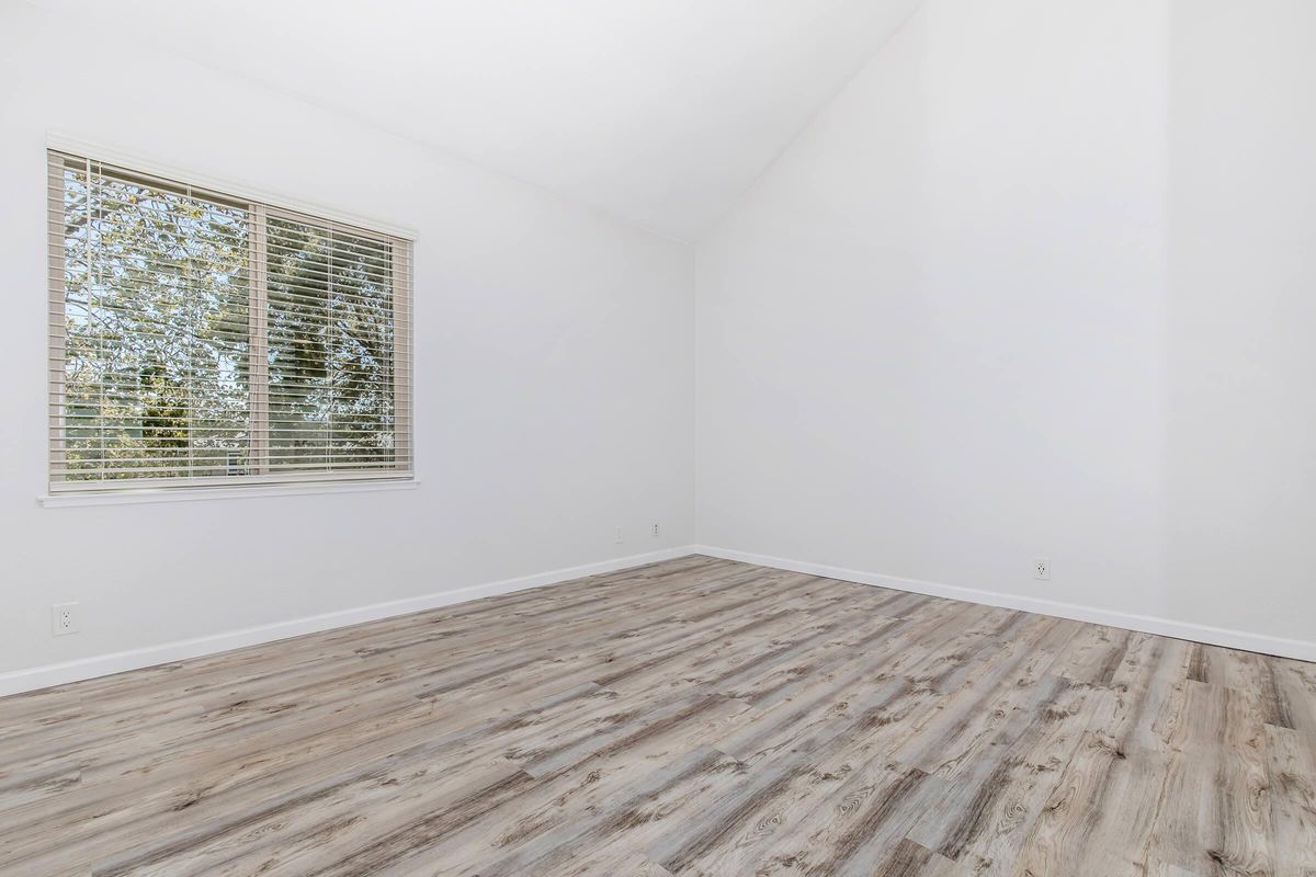 Empty room with white walls, a high ceiling, and a large window letting in natural light. The floor is covered with light wood laminate, and there are no furnishings present. The window features blinds, and outside greenery is visible through the glass.