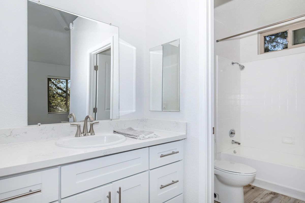 Modern bathroom with a dual sink vanity featuring a white countertop and ample storage. A large mirror above the sinks reflects the bright, airy space. The bathroom includes a shower stall with white walls and a toilet, set against light wood-style flooring. Natural light filters in through a window.