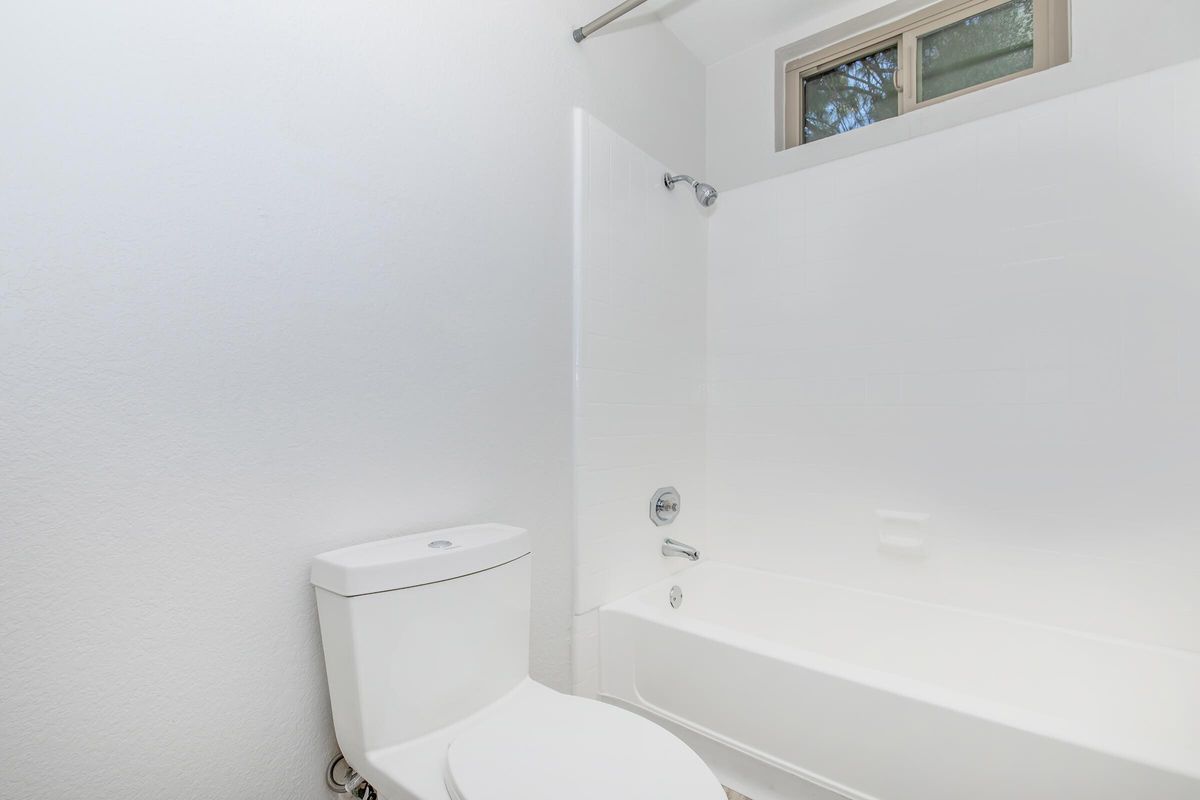 A clean, modern bathroom featuring a white bathtub, showerhead, and a toilet. The walls are painted white, and there is a window above the bathtub allowing natural light to enter the space. The overall design is simple and minimalistic.