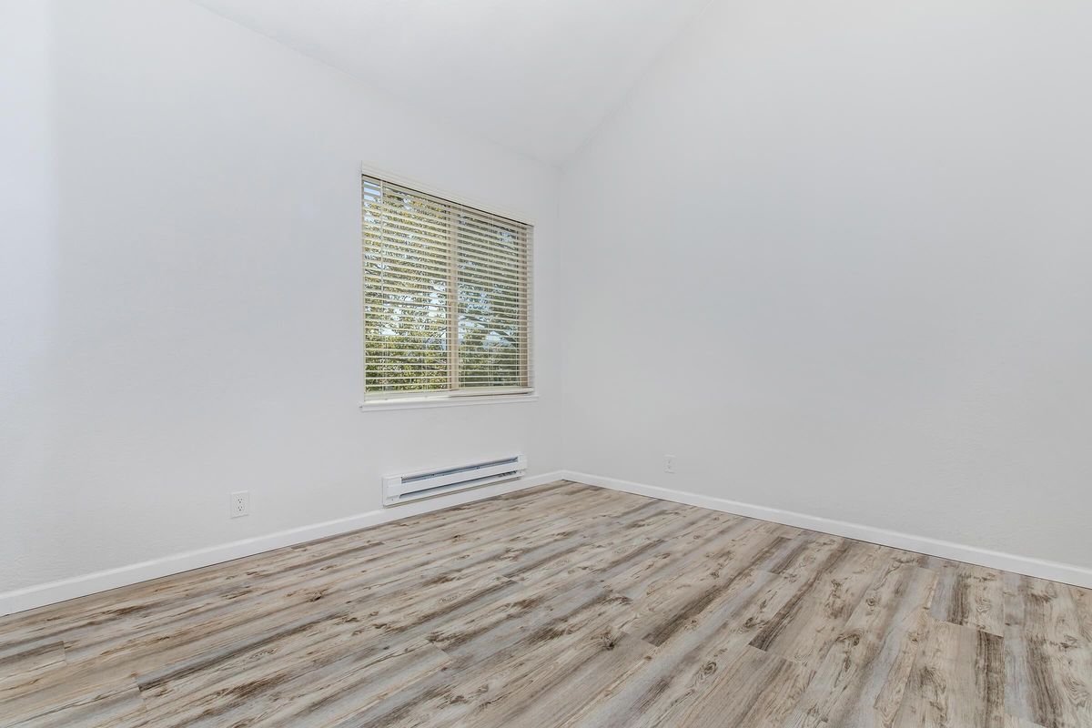 A bright, empty room with white walls and a high ceiling. There is a large window with blinds, allowing natural light to enter. The floor is covered in light-colored wooden laminate, creating a clean and spacious atmosphere. A heating vent is visible on the wall beneath the window.