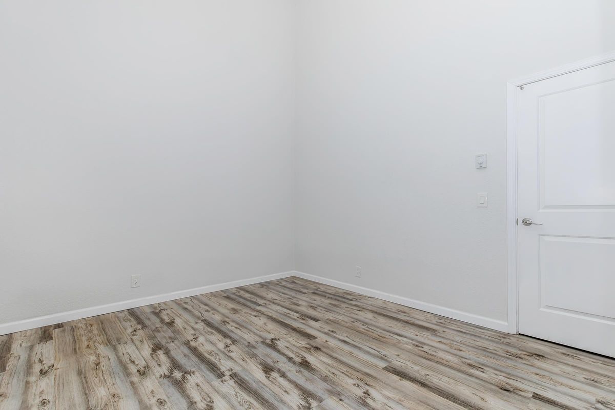 Empty room with light gray walls and a wooden floor. The space features a single white door on the right side, and there are no furnishings or decorations present, creating a minimalist and spacious appearance. Natural light may be entering, contributing to the bright atmosphere.