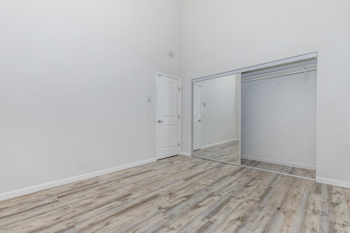A spacious, empty room featuring light-colored walls and a high ceiling. The wooden floor has a warm tone, and there is a large mirror reflecting a closet area. A white door is visible on the left side of the image, enhancing the room's modern and minimalist aesthetic.