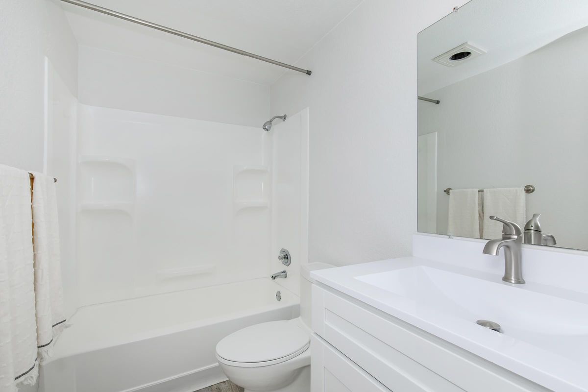 A clean, modern bathroom featuring a white bathtub with a shower, a white toilet, and a sleek vanity with a sink. The walls are painted in a light color, and there is a large mirror above the sink. Towels are neatly hung on a rack, contributing to the tidy and airy atmosphere of the space.