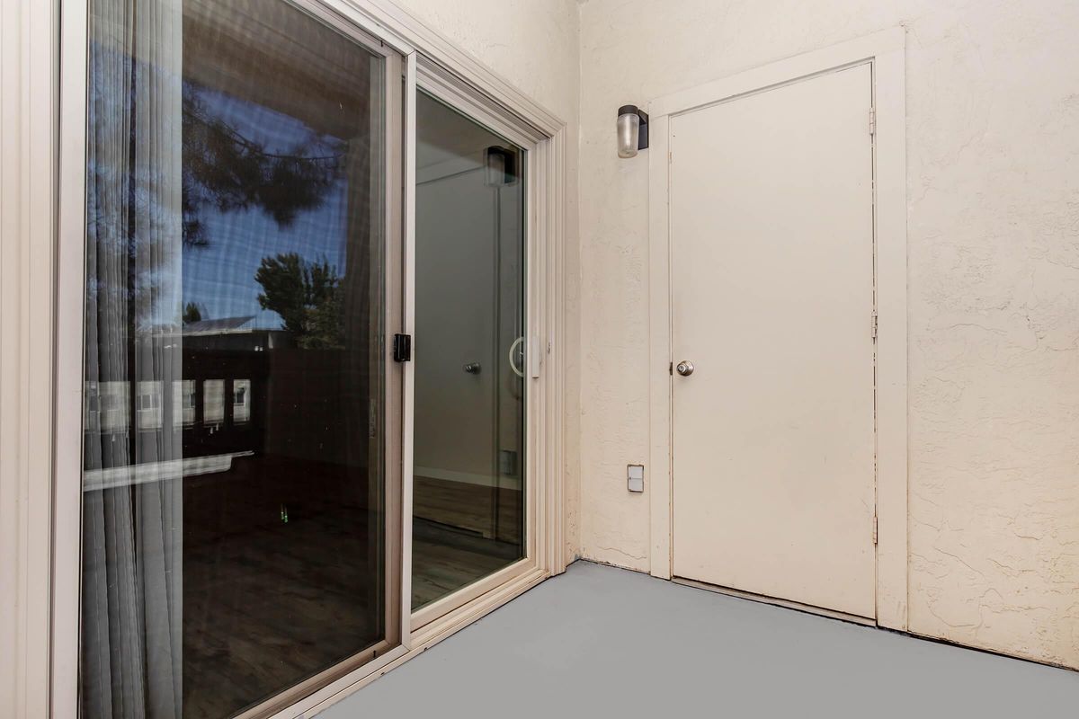 A light-colored, textured wall with a sliding glass door leading to an outdoor area. The door has sheer curtains, and a white door is visible on the right side. The floor is painted gray, creating a simple and clean look. Natural light filters in, giving a bright appearance.