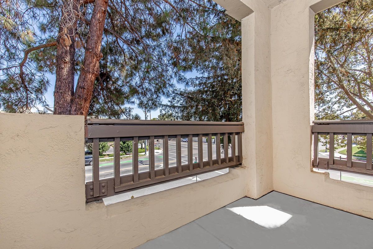 View from a balcony displaying a corner perspective with a concrete floor and beige walls. A wooden railing separates the balcony from an outdoor scene featuring trees and a street visible in the background under a clear sky. Natural light casts shadows on the floor.