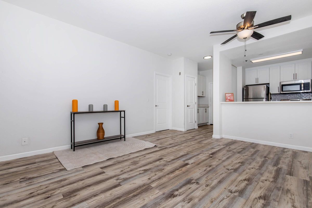 A modern, minimally furnished living space featuring light wood flooring. A small black table with decorative vases in orange and gray colors is positioned against a white wall. The background shows an open kitchen area with stainless steel appliances and white cabinetry. Soft beige rug on the floor adds warmth to the room.