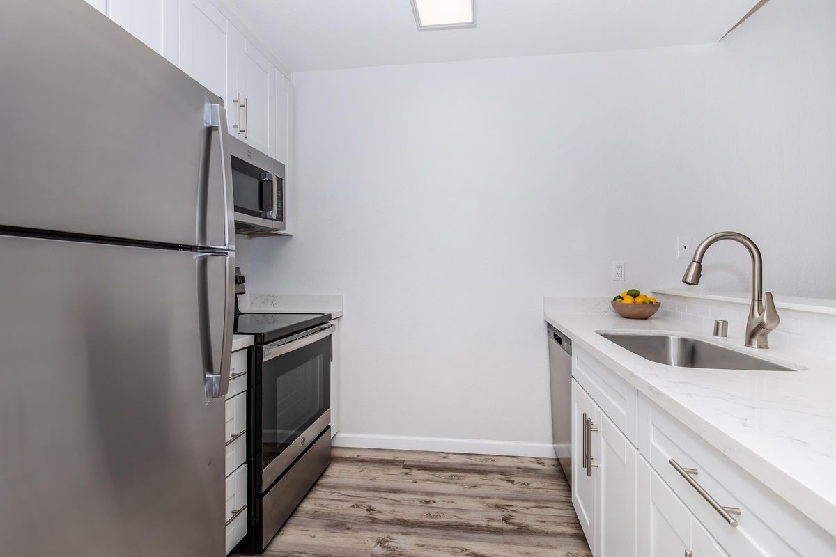 A modern kitchen featuring stainless steel appliances, including a refrigerator, microwave, and oven. The space has white cabinets, a marble countertop, and a large sink. Wood-like flooring adds warmth to the area, while a bowl of fruit sits on the countertop, adding a touch of color.