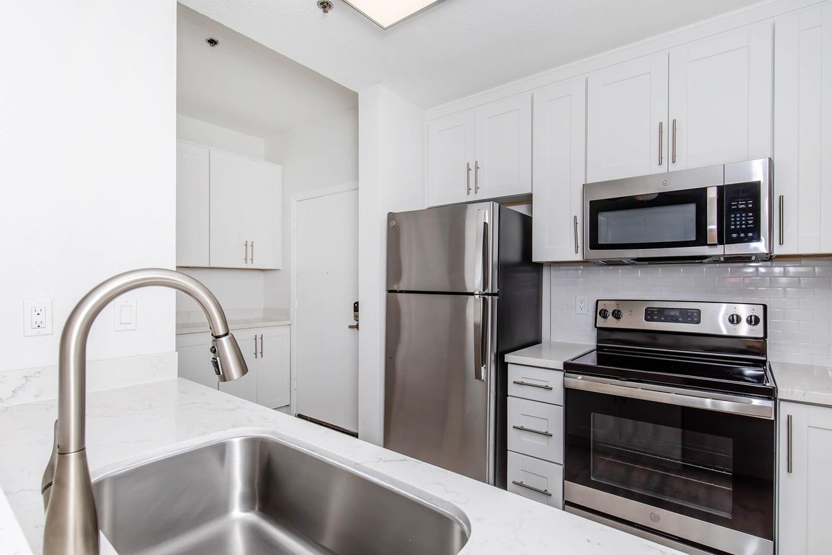Modern kitchen featuring stainless steel appliances, including a refrigerator, microwave, and oven. The cabinets are white with sleek handles, and the countertop is made of light-colored stone. A stainless steel sink is visible in the foreground, with a view towards a door leading to another area.