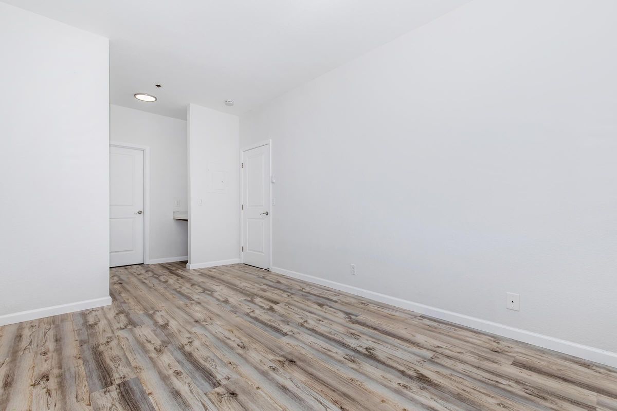 A spacious, empty room featuring light-colored walls and a wooden floor. A closed door is visible on the left leading to another space, while a second door on the right appears to lead to a bathroom or closet. The overall ambiance is bright and airy, with ample natural light.