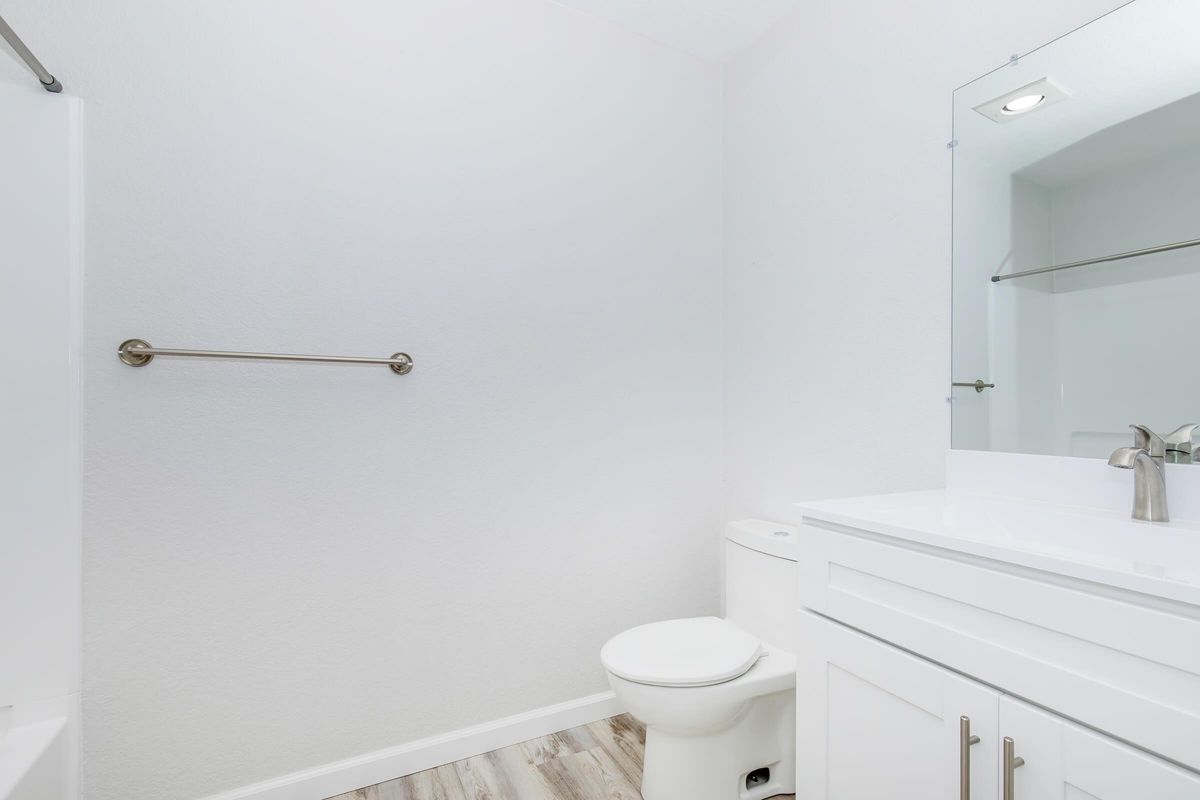 A clean, modern bathroom featuring a white toilet and a sleek vanity with a sink. The walls are painted a light color, and there is a large mirror above the sink. A towel bar is mounted on the wall, and the floor has a light wooden texture, creating a bright and spacious atmosphere.