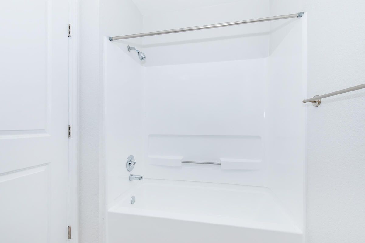 A clean, modern bathroom featuring a white tub with a shower curtain rod, minimalist design, and smooth white walls. The space appears well-lit and spacious, with a door visible in the background.