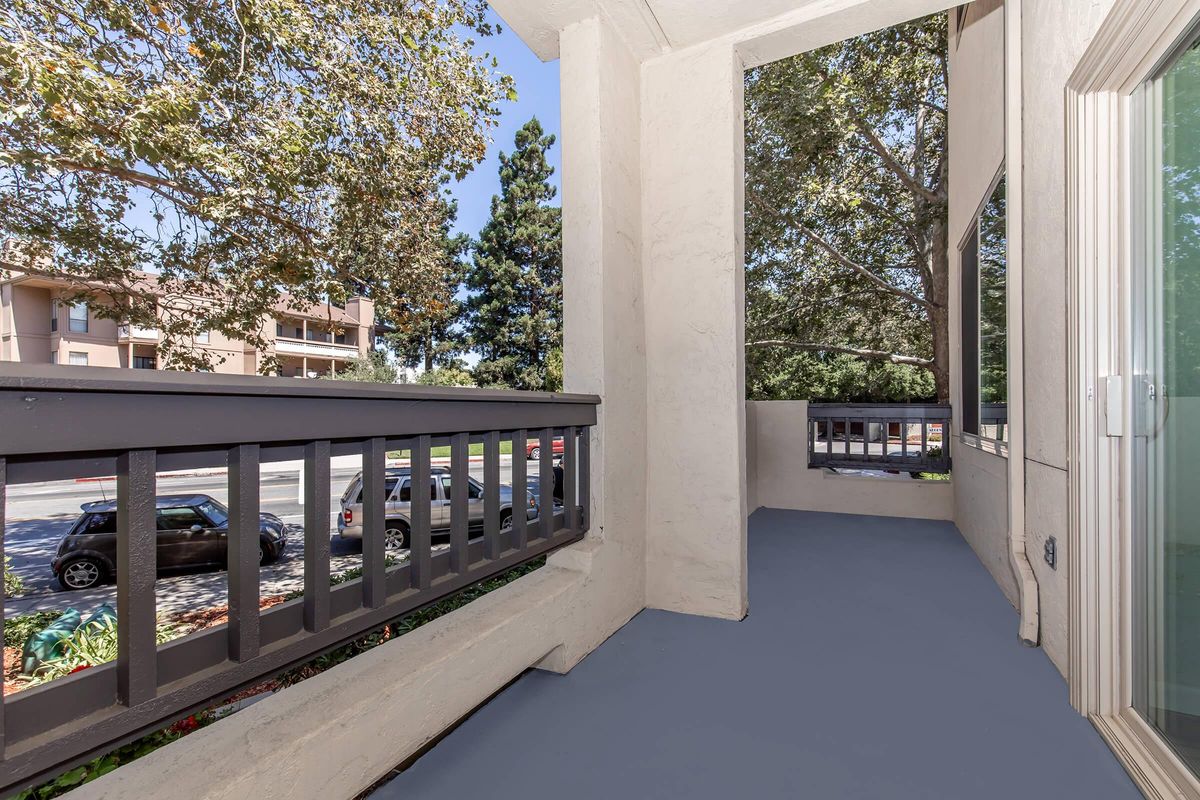 A view from a balcony featuring a gray railing and light-colored walls. The floor is painted blue, and there are trees outside providing shade. In the background, parked cars and a multi-story building are visible, under a clear blue sky.