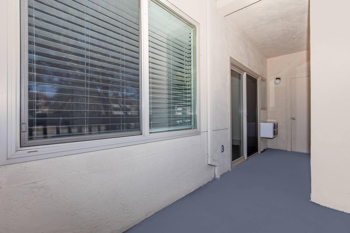 A view of a light-colored hallway with large windows featuring horizontal blinds. On the left, a window allows natural light to enter, while on the right, there are glass doors leading to an outdoor area. The floor is painted in a muted color, and the walls are smooth and textured.
