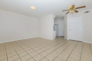 Spacious, empty room with beige walls and tiled floor. A ceiling fan is installed, and there is a light fixture on the ceiling. A doorway leads to a kitchen area, and a closed door is visible to the right. The room is well-lit, creating a welcoming atmosphere.