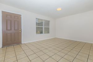 A spacious, empty room featuring light-colored walls and beige tiled flooring. The room includes a front door on the left with a simple design and a window with blinds opposite the door, allowing natural light to enter. The overall ambiance is bright and neutral, suitable for various decorating styles.