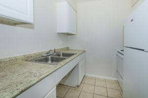 A small, tidy kitchen featuring a double sink with a polished countertop, white cabinets above, and a white refrigerator on the right. The walls are painted a light color, and the floor is tiled with cream-colored tiles. There is an oven on the far right, completing the compact kitchen layout.