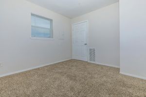 Empty room with light beige carpet, featuring a white wall with a window and a door. The room is sparsely decorated, emphasizing a clean and minimalistic look, with a small air vent on the floor. Natural light enters through the window, illuminating the space.
